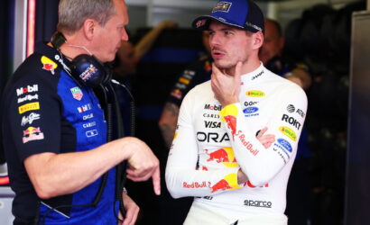 Two Formula 1 crew members chat in a garage, one in a white Red Bull racing suit and cap, the other in a blue team shirt with headphones nearby.