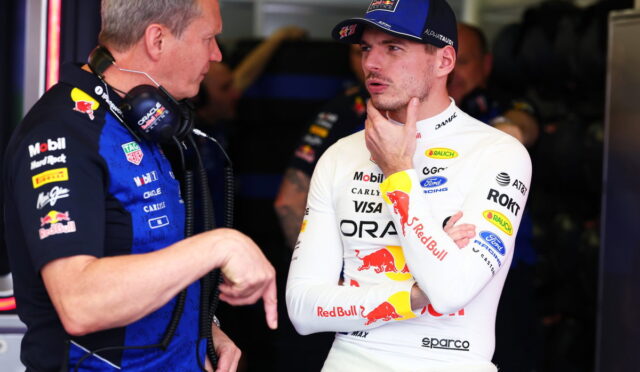 Two Formula 1 crew members chat in a garage, one in a white Red Bull racing suit and cap, the other in a blue team shirt with headphones nearby.