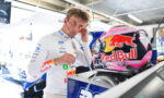 Driver in a white Red Bull racing suit adjusts a headset while standing next to a pink Red Bull helmet in a garage pit area.