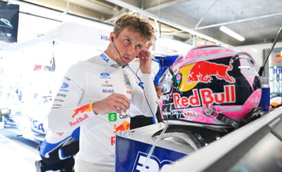 Driver in a white Red Bull racing suit adjusts a headset while standing next to a pink Red Bull helmet in a garage pit area.