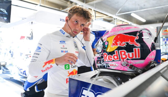 Driver in a white Red Bull racing suit adjusts a headset while standing next to a pink Red Bull helmet in a garage pit area.