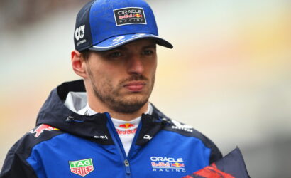 Male race car driver in blue Oracle Red Bull Racing jacket and cap, looking off to the side in a soft, outdoor setting.