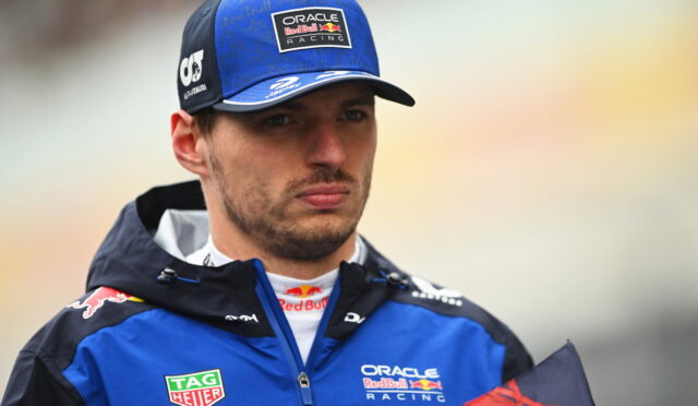 Male race car driver in blue Oracle Red Bull Racing jacket and cap, looking off to the side in a soft, outdoor setting.