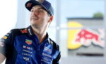 Male race car driver in a blue Red Bull racing polo and cap, looking up, with sponsor logos visible.