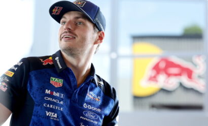 Male race car driver in a blue Red Bull racing polo and cap, looking up, with sponsor logos visible.