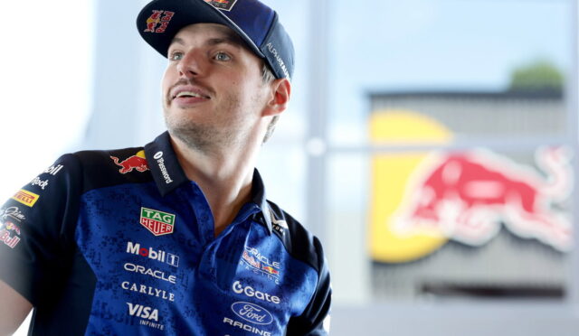 Male race car driver in a blue Red Bull racing polo and cap, looking up, with sponsor logos visible.