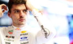 Male racecar driver in a white racing suit adjusts his hair in a garage, sponsor logos visible.