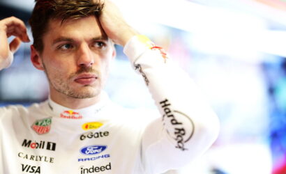 Male racecar driver in a white racing suit adjusts his hair in a garage, sponsor logos visible.