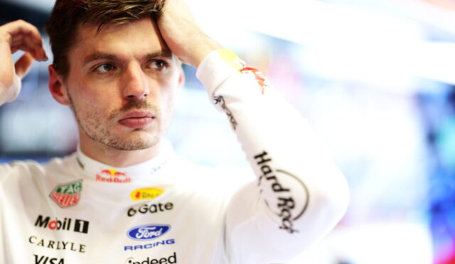 Male racecar driver in a white racing suit adjusts his hair in a garage, sponsor logos visible.
