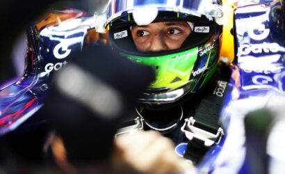Close-up of a race car driver wearing a blue-green helmet, eyes visible through the visor, strapped into the cockpit.