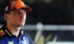 Portrait of a male race car driver wearing an orange cap with sponsor logos and a blue team shirt with patches.