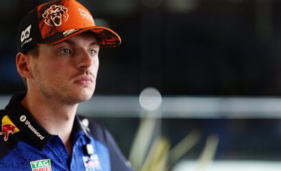 Portrait of a male race car driver wearing an orange cap with sponsor logos and a blue team shirt with patches.