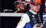 Portrait of a Formula 1 driver in a sponsor-laden blue and white racing suit, wearing a cap, in a pit garage with a helmet visible in the background.
