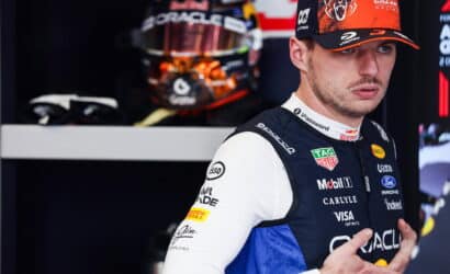 Portrait of a Formula 1 driver in a sponsor-laden blue and white racing suit, wearing a cap, in a pit garage with a helmet visible in the background.
