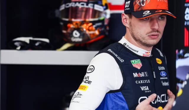 Portrait of a Formula 1 driver in a sponsor-laden blue and white racing suit, wearing a cap, in a pit garage with a helmet visible in the background.