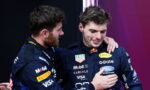 Two race car drivers in blue sponsor suits share a congratulatory hug on stage.