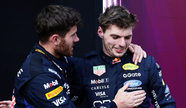 Two race car drivers in blue sponsor suits share a congratulatory hug on stage.