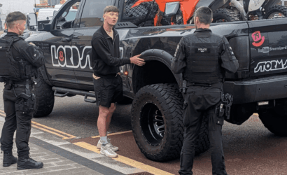 Young man in a black zip-up jacket speaks to two police officers in tactical gear beside a lifted black pickup with bikes in the bed.