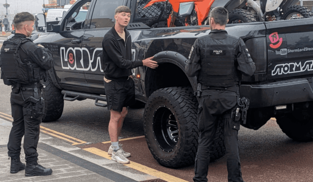 Young man in a black zip-up jacket speaks to two police officers in tactical gear beside a lifted black pickup with bikes in the bed.