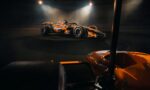 Orange Formula 1 race car displayed on a circular platform under bright spotlights in a dark showroom/studio set-up