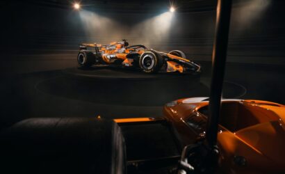 Orange Formula 1 race car displayed on a circular platform under bright spotlights in a dark showroom/studio set-up