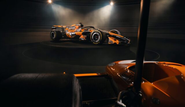 Orange Formula 1 race car displayed on a circular platform under bright spotlights in a dark showroom/studio set-up