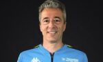 Portrait of a man in a blue Trackhouse MotoGP Team jersey against a dark background, smiling at the camera.
