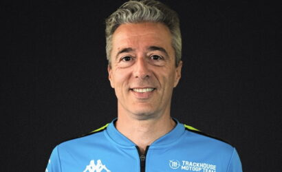Portrait of a man in a blue Trackhouse MotoGP Team jersey against a dark background, smiling at the camera.