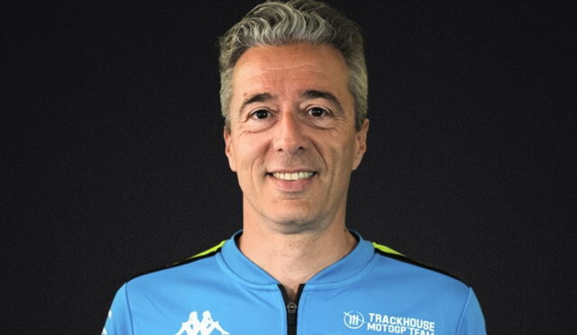 Portrait of a man in a blue Trackhouse MotoGP Team jersey against a dark background, smiling at the camera.