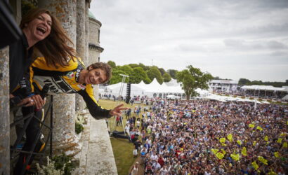 Two people lean from a stone balcony, laughing and waving to a crowded outdoor festival with white tents and a stage in the background.