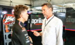Racer in a black suit talks with an older man in a white shirt beside an Aprilia Racing backdrop in a workshop.
