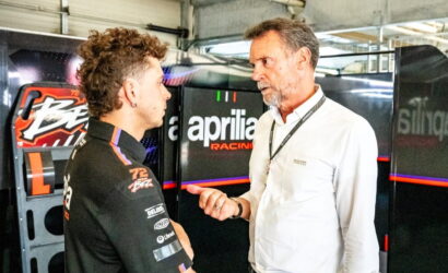 Racer in a black suit talks with an older man in a white shirt beside an Aprilia Racing backdrop in a workshop.