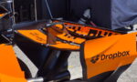 Orange race bike with carbon fiber components on a stand in a pit area, Dropbox branding on the side panel.