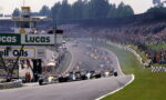 Formula 1 cars at the start on a wide track with sponsor banners and a packed grandstand in the background.