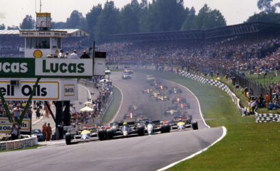 Formula 1 cars at the start on a wide track with sponsor banners and a packed grandstand in the background.