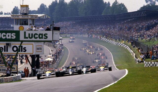 Formula 1 cars at the start on a wide track with sponsor banners and a packed grandstand in the background.
