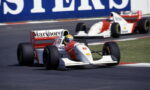 Two vintage Formula One cars on a race track, white with red Marlboro livery, chasing into a corner with a blue sponsor wall in the background.