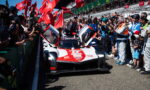 Race car with doors open, crowds cheering on both sides at a pit-lane moment before a race start, red flags waving nearby.