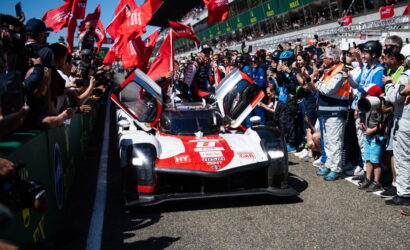 Race car with doors open, crowds cheering on both sides at a pit-lane moment before a race start, red flags waving nearby.