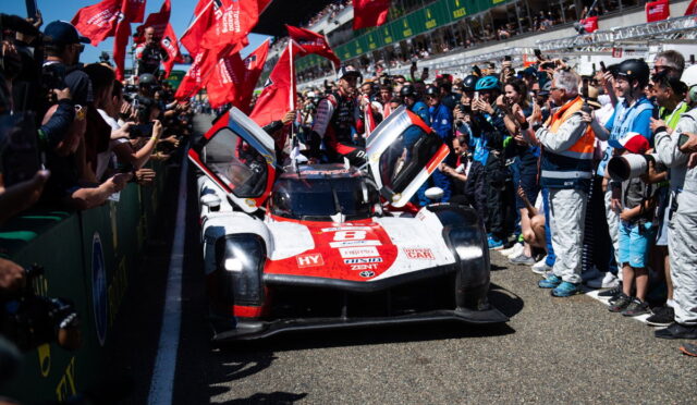 Race car with doors open, crowds cheering on both sides at a pit-lane moment before a race start, red flags waving nearby.