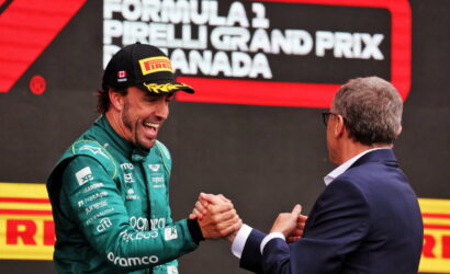 Formula 1 driver in green suit and Pirelli cap shakes hands with a man in a suit on a podium, smiling after a race win.