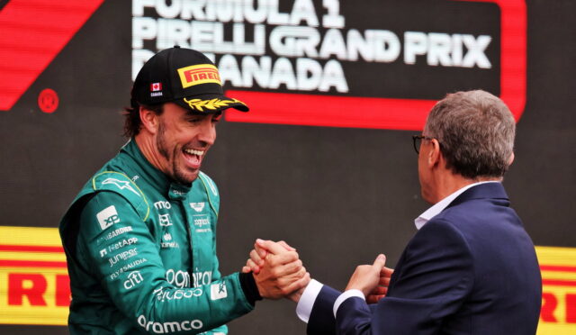 Formula 1 driver in green suit and Pirelli cap shakes hands with a man in a suit on a podium, smiling after a race win.