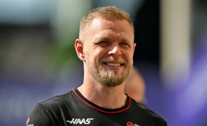 Smiling man with short blond hair and beard, wearing a black athletic shirt with red trim and sponsor logos.