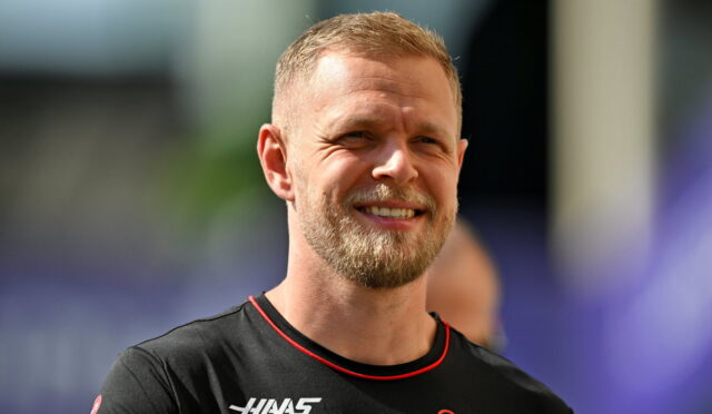 Smiling man with short blond hair and beard, wearing a black athletic shirt with red trim and sponsor logos.