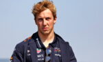 Young racing driver with blond hair, navy jacket and sunglasses hanging from the collar, facing the camera.