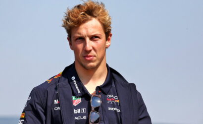 Young racing driver with blond hair, navy jacket and sunglasses hanging from the collar, facing the camera.