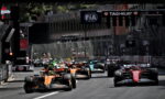 Group of Formula 1 cars sprinting off the start on a city street circuit, orange car leading the pack under sponsor banners.