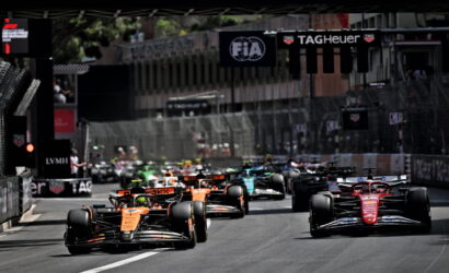 Group of Formula 1 cars sprinting off the start on a city street circuit, orange car leading the pack under sponsor banners.