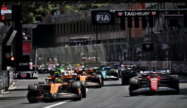 Group of Formula 1 cars sprinting off the start on a city street circuit, orange car leading the pack under sponsor banners.