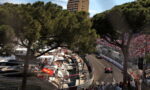 Race cars navigate a tight harbor street circuit as spectators fill grandstands; boats docked at left behind pine trees.